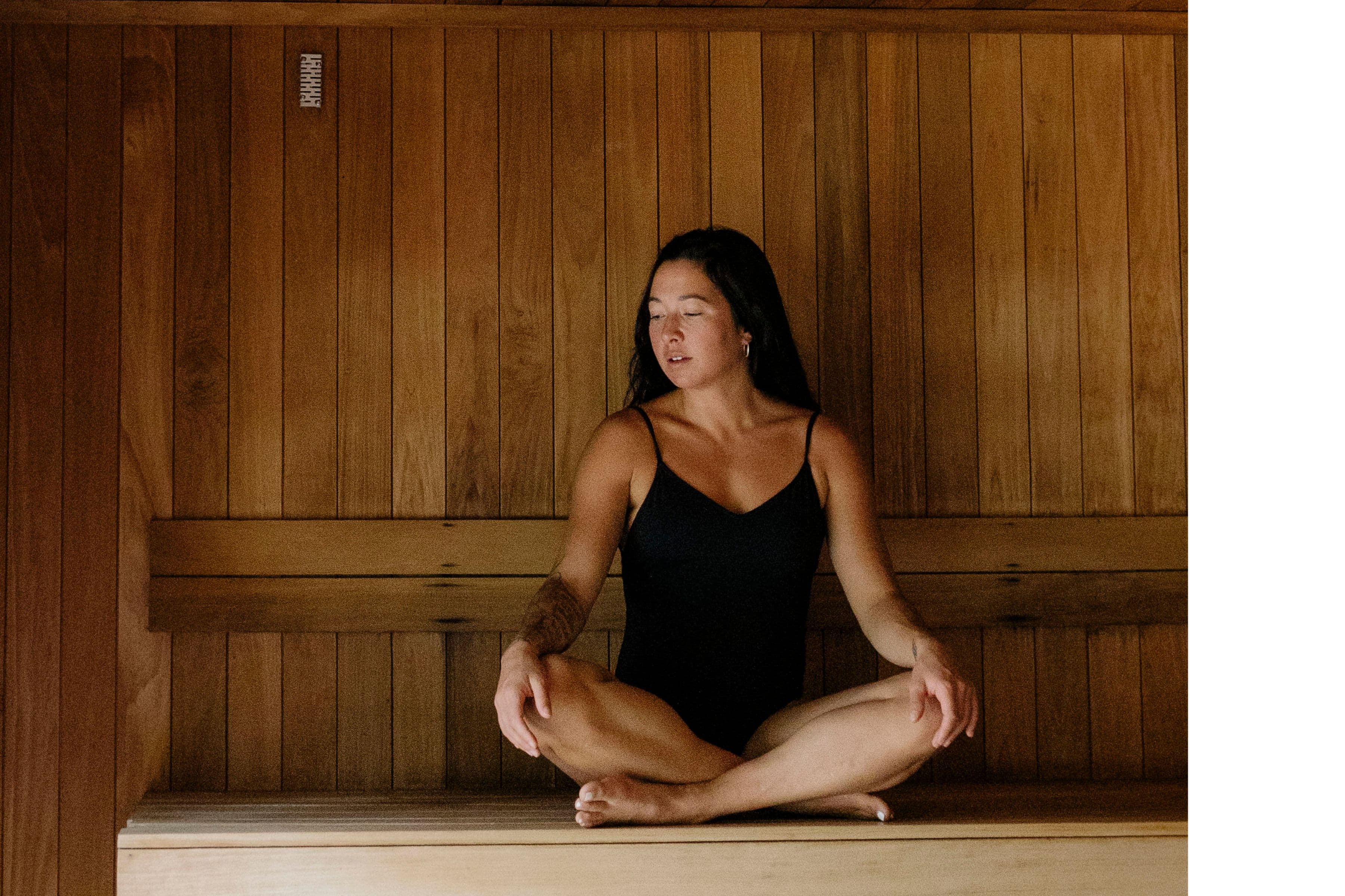 A woman sitting cross-legged in a wooden sauna, wearing a black swimsuit, with her eyes closed in a relaxed pose. The wooden panels create a warm atmosphere, perfect for a unique thermal experience.