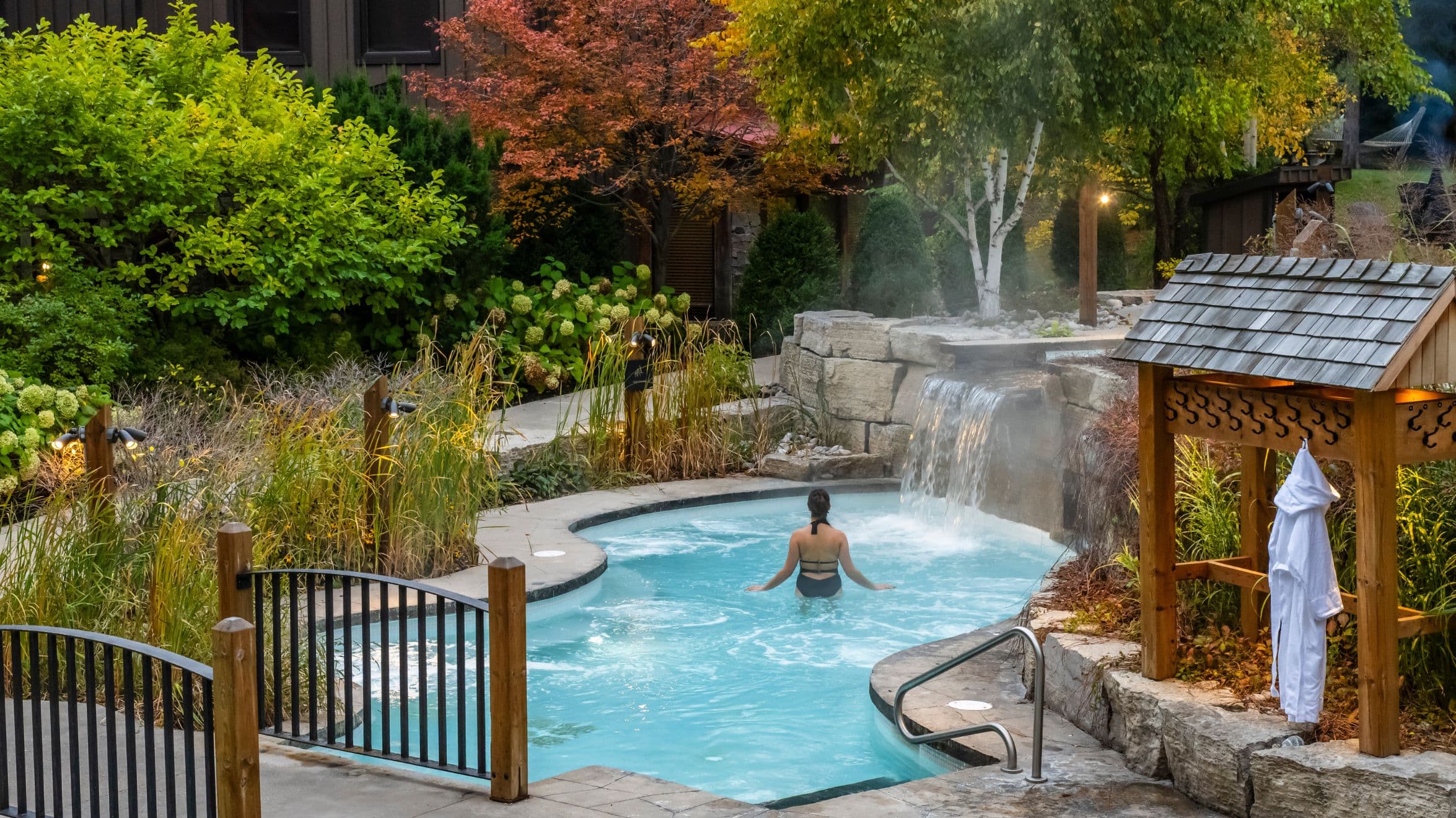 A person relaxes in a small outdoor pool surrounded by lush greenery and trees with autumn leaves.