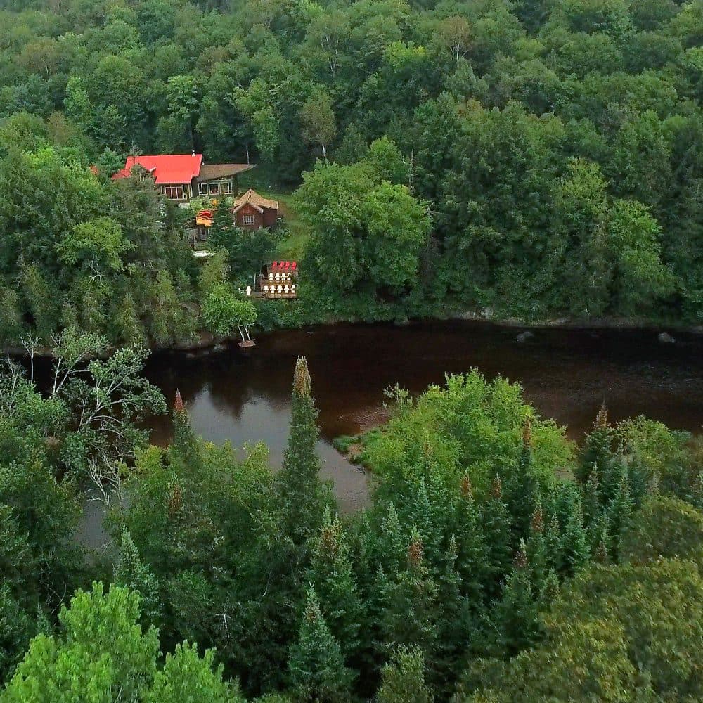 Aerial view of a lush forest with a river running through it. Nestled among the trees is a house with a bright red roof and wooden exterior.