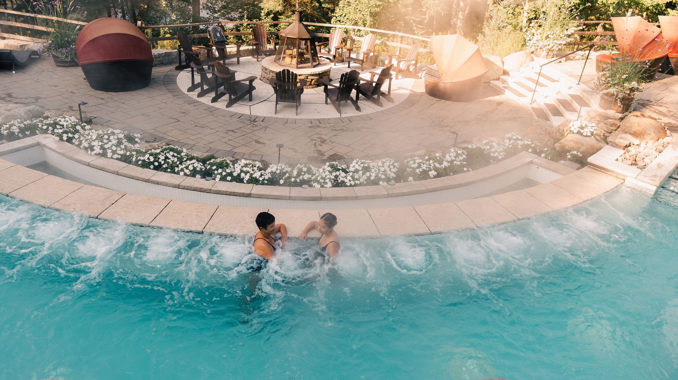 Two people relax in a curved pool with jets, surrounded by greenery and a patio area with chairs and tables.